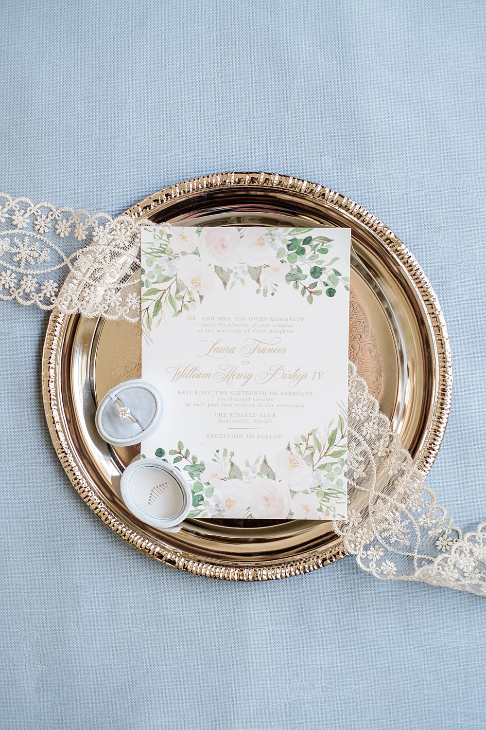 Wedding invitation and ring sit atop a silver tray on a dusty blue background