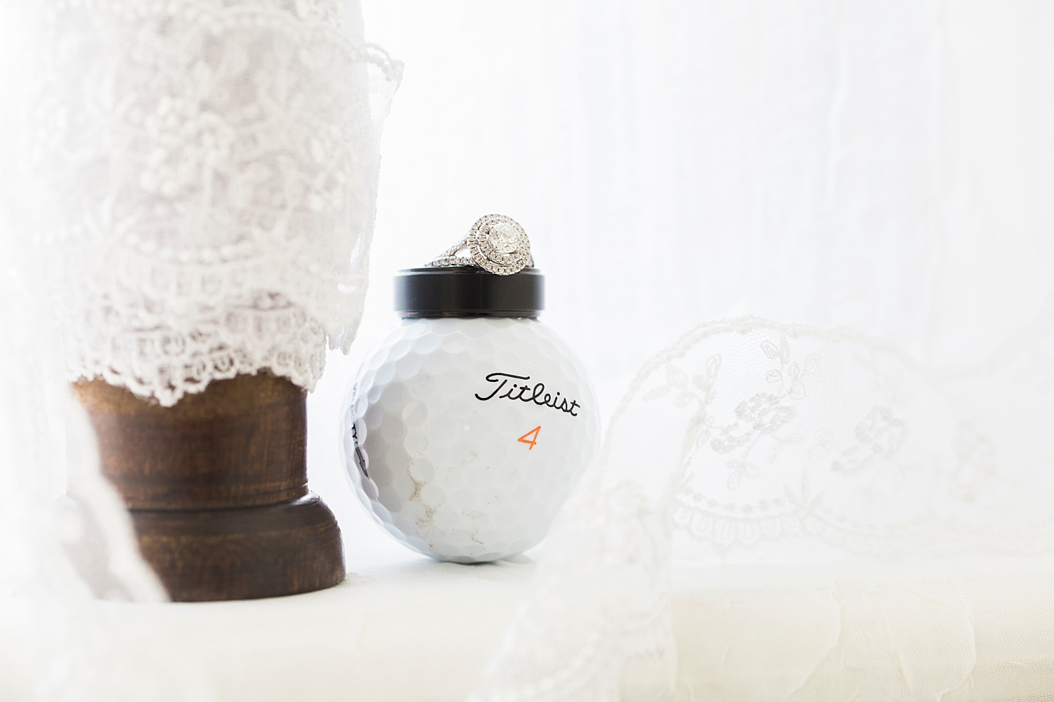 Close up photo of wedding rings sitting on a golf ball behind flowing white lace