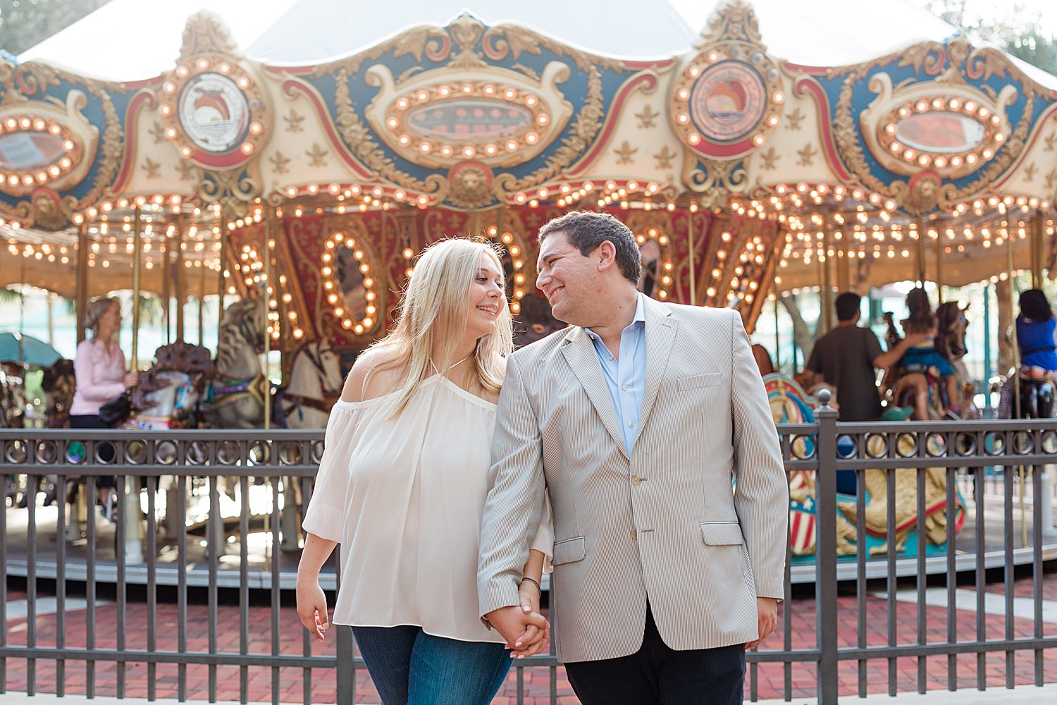 Engaged couple holds hands and smiles in front of a carousel