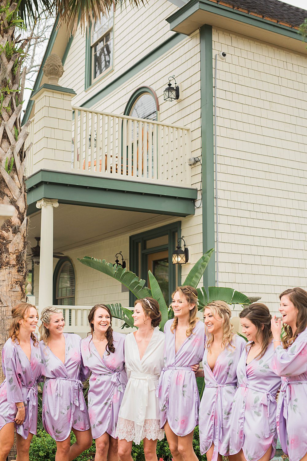 Bride and her bridesmaids laugh while standing in matching lavender robes outside of the Collector Inn